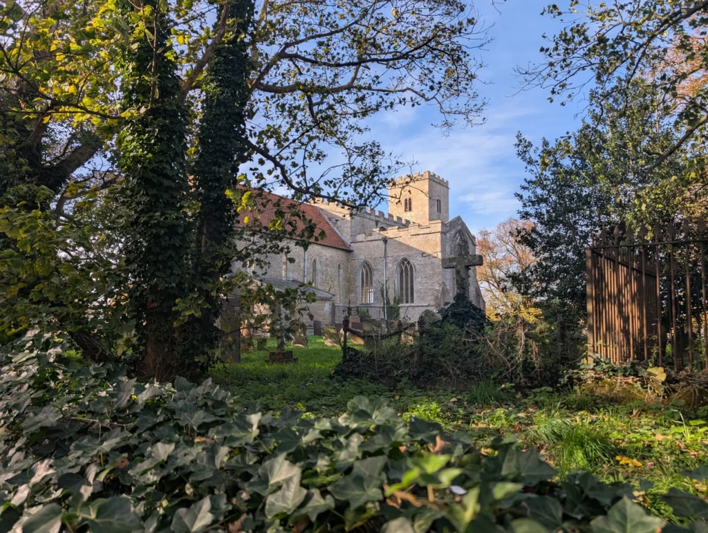 A photo of a stone church, with trees, graves and an ivy covered wall in the foreground.